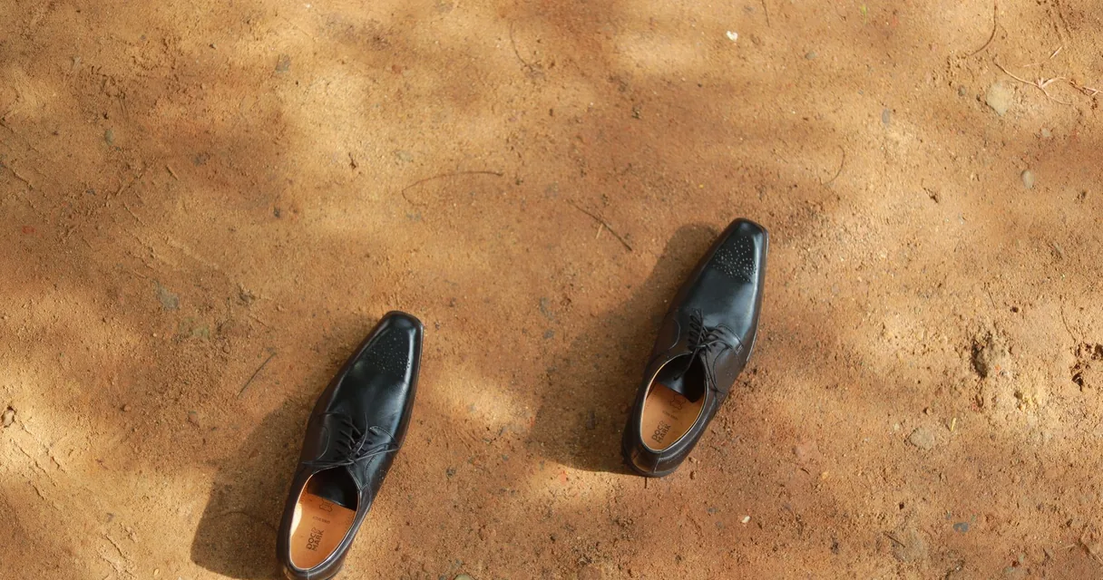 Par de sapatos sociais pretos sobre chão de terra, ilustrando o dedo do pé doendo ao calçar sapato