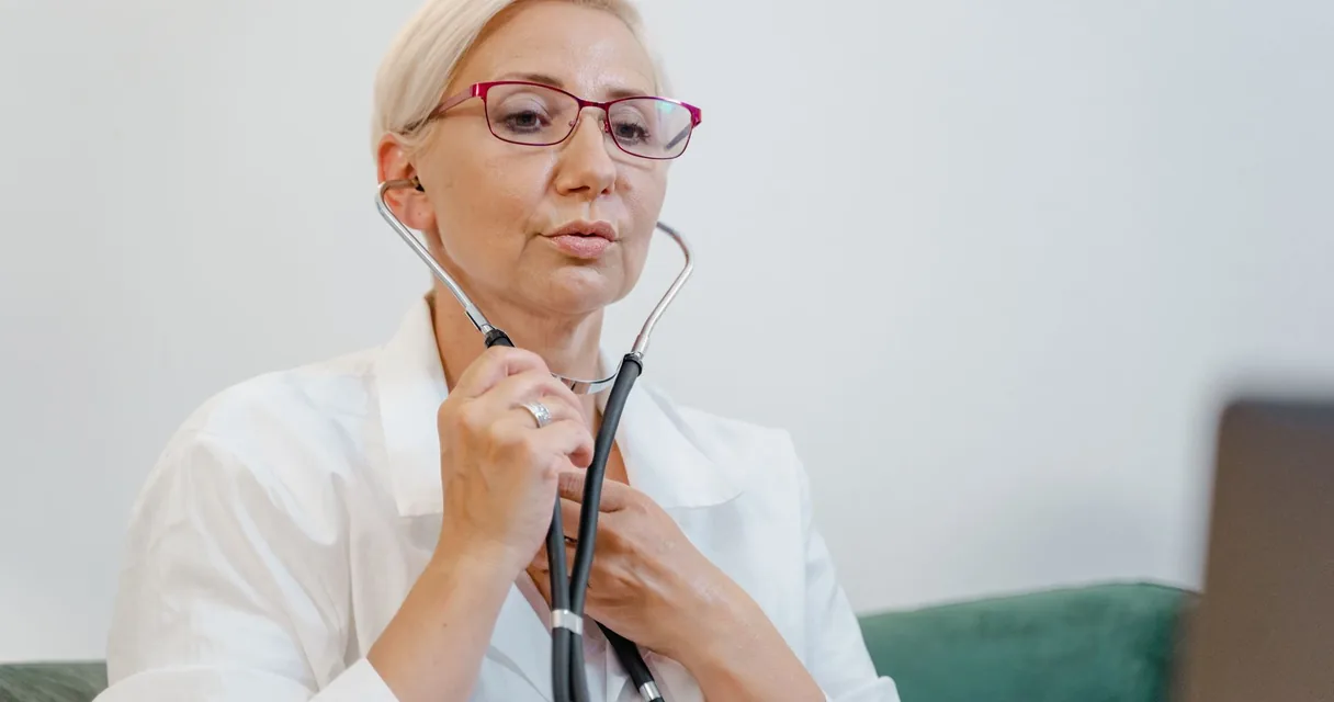 Médica de cabelo grisalho e óculos vermelhos colocando o estetoscópio nos ouvidos em consultório